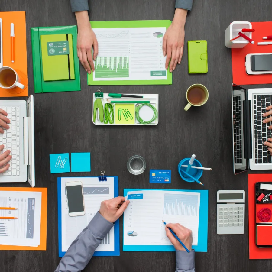 Top view of a workspace with laptops, documents, smartphones, coffee cups, and office supplies arranged neatly on a dark table.