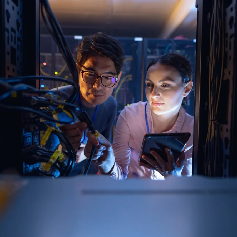 Two IT technicians work together on server cables, troubleshooting and using a tablet in a dim data center.