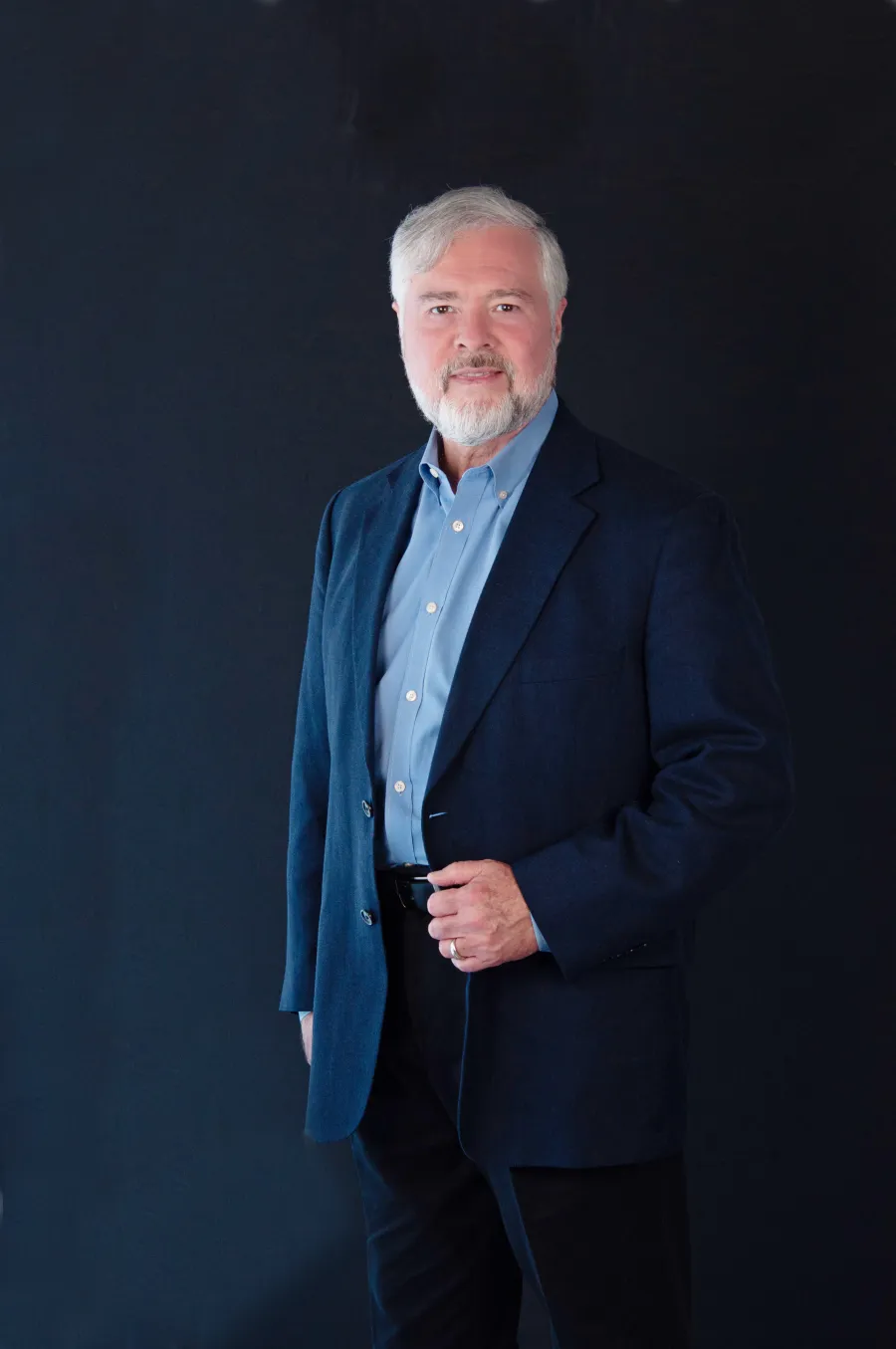 Mature man with gray hair and beard wearing a blue blazer and light blue shirt against a dark background.