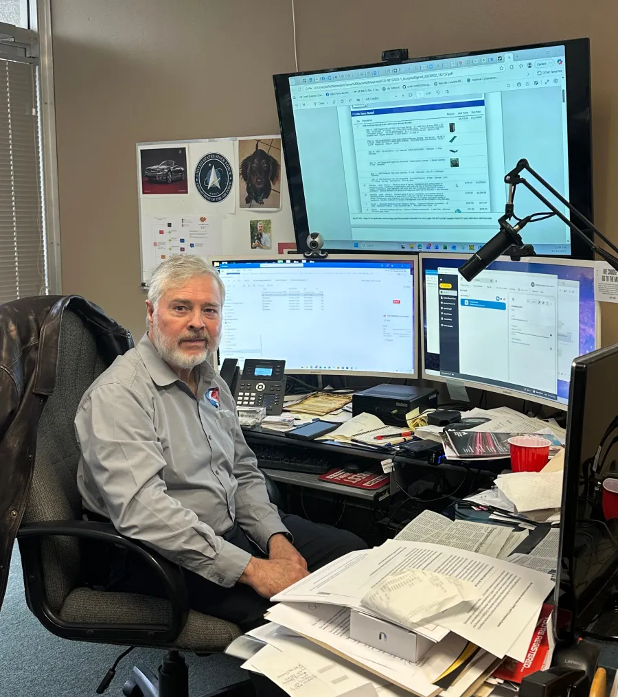 Man with gray hair sitting at cluttered desk with multiple computer monitors and office equipment in workspace