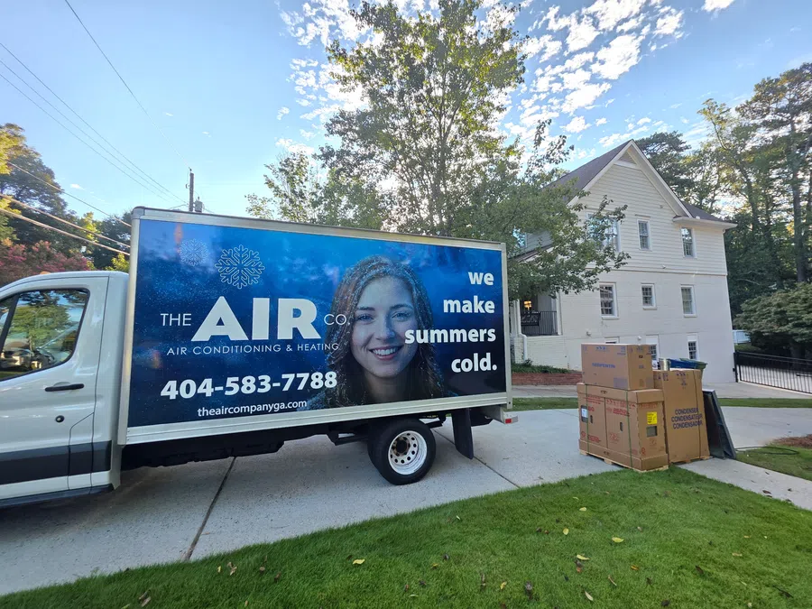 Air conditioning company truck with smiling woman and text we make summers cold parked near home with AC unit boxes.