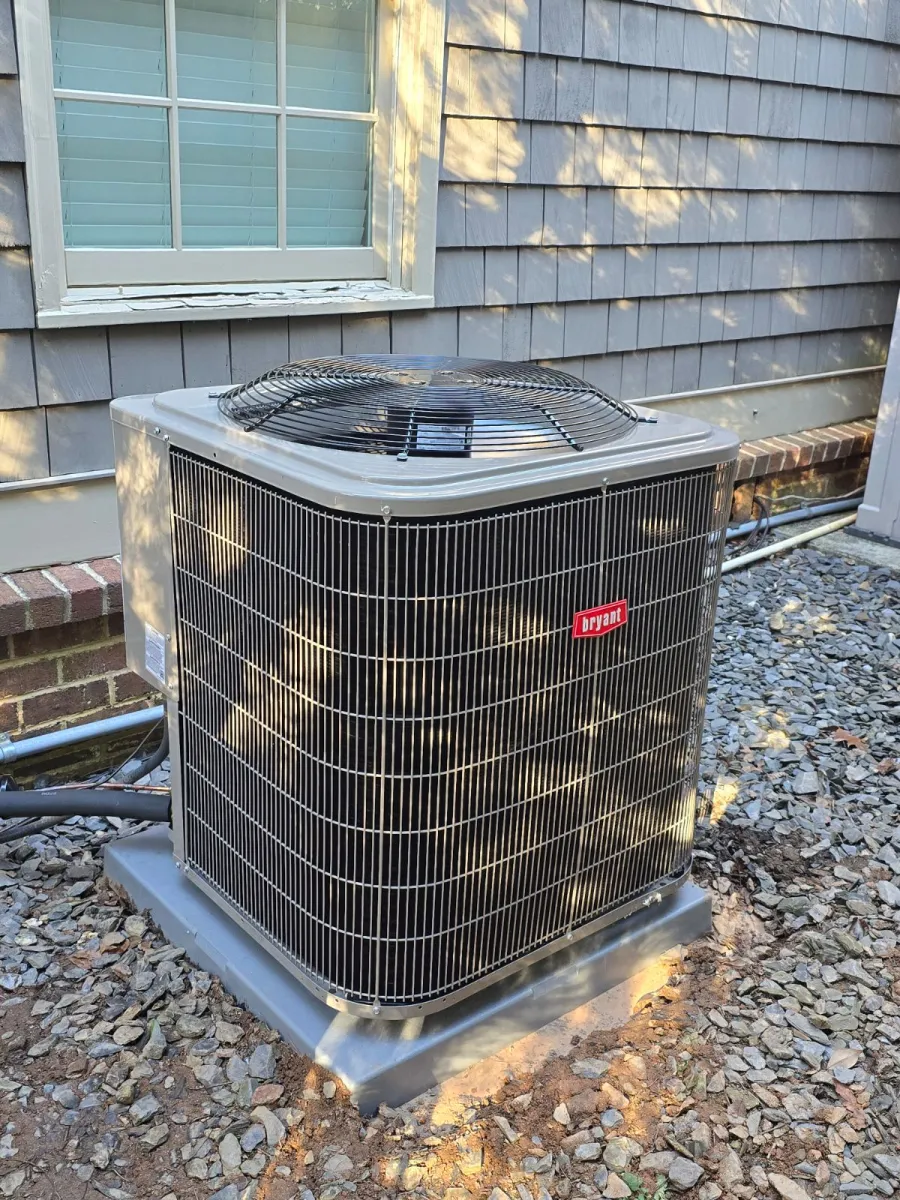 Outdoor Bryant air conditioning unit installed on a concrete pad beside a house with gray siding and gravel ground.