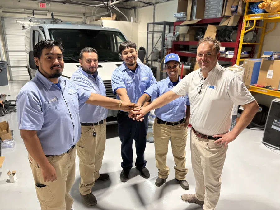 Team of five HVAC technicians in uniform joining hands inside a workshop with a white service van in the background