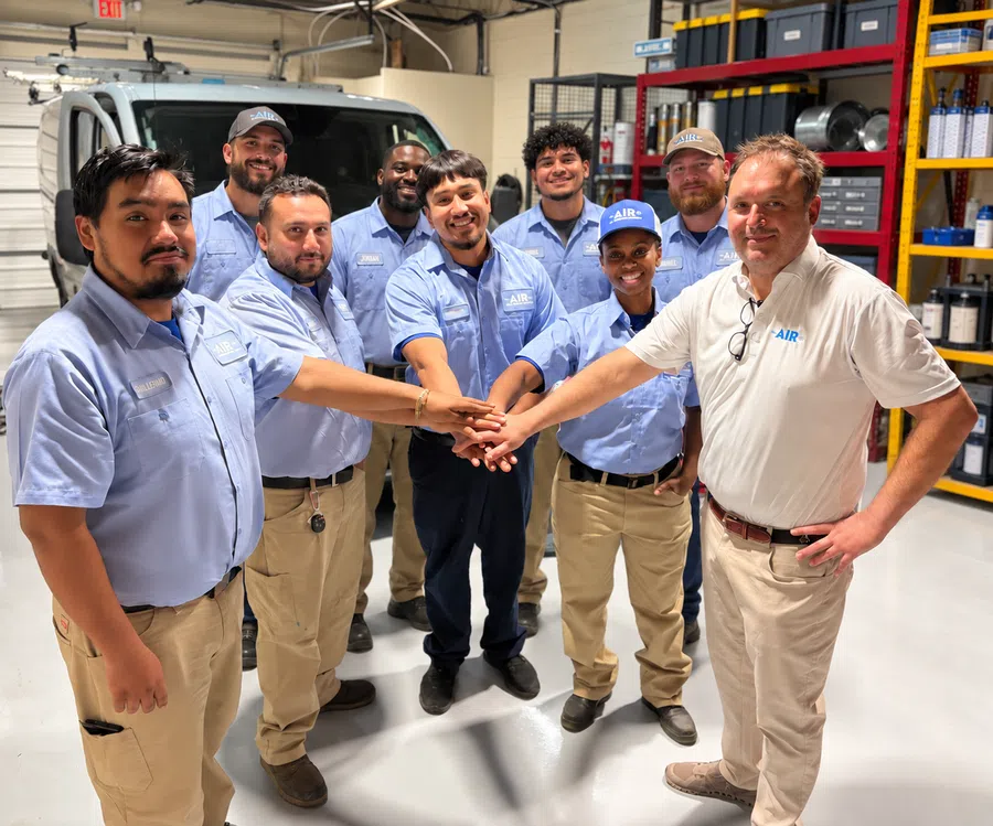 Diverse team of HVAC technicians in uniform joining hands in a workshop with tools and van in background.