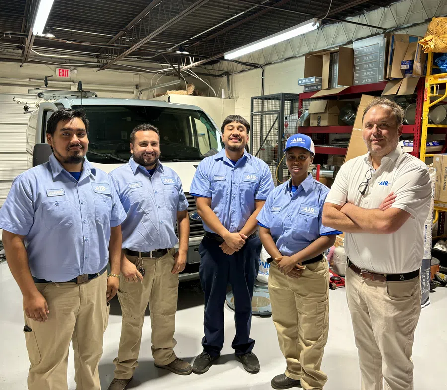 Five HVAC technicians standing in a workshop wearing company uniforms with a service van in the background.