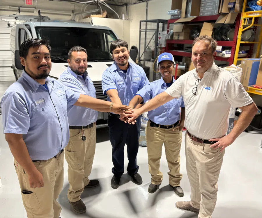 Team of five HVAC technicians in uniform joining hands inside workshop with equipment and van in background.