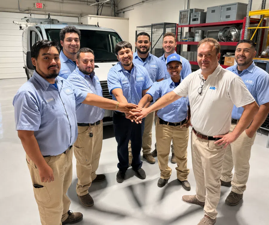 A group of HVAC technicians in blue uniforms join hands in a warehouse with service vehicles and equipment.