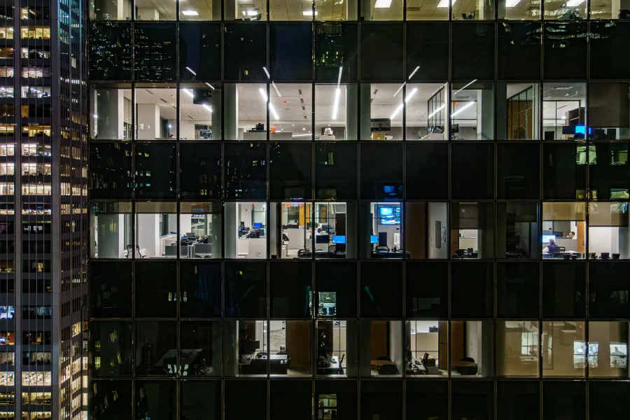 Night view of illuminated office spaces inside a modern glass skyscraper with visible desks and computers.
