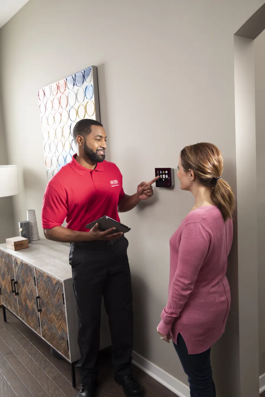 Technician in red shirt demonstrating home security touchscreen panel to woman in pink sweater inside a modern room