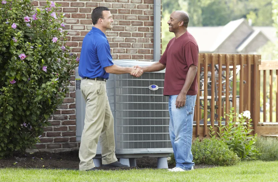 Technician and homeowner shaking hands next to an air conditioning unit outside a brick house on a sunny day.