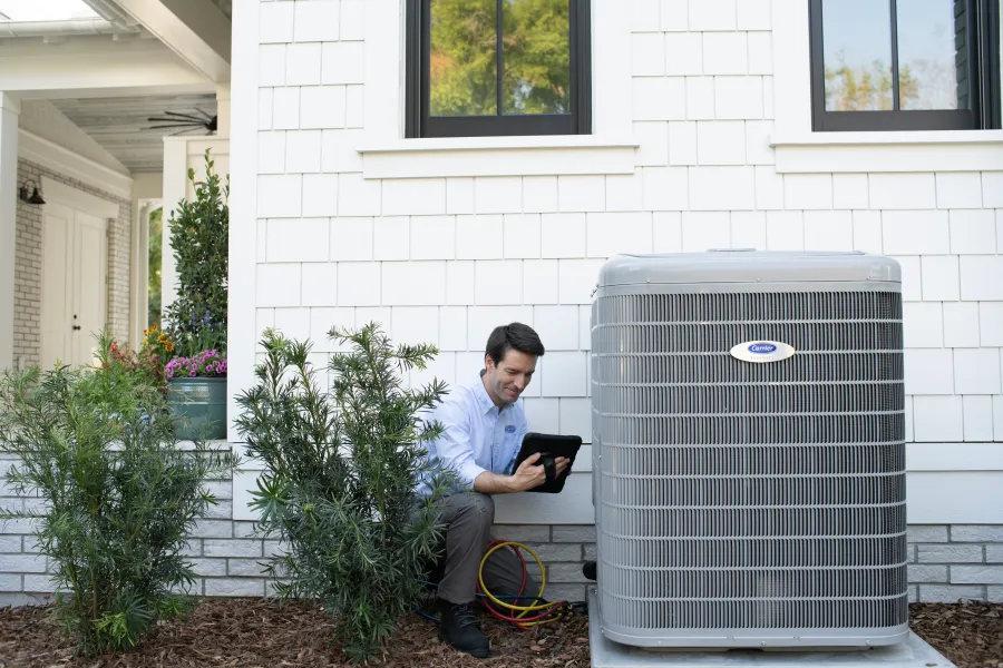 Technician inspecting a large Carrier air conditioning unit outside a white house using a tablet device.