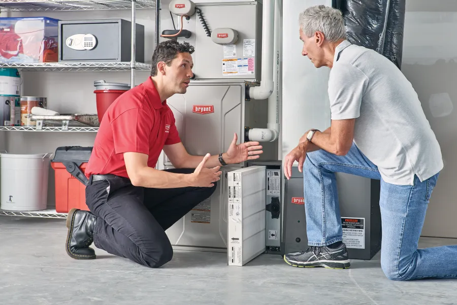 Technician in red shirt explains HVAC system to a customer in a utility room with shelves and equipment.