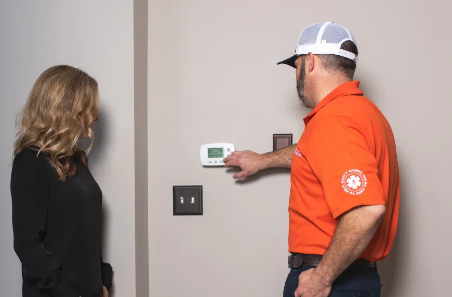 Technician in orange shirt adjusting a thermostat on a beige wall while woman watches closely indoors.