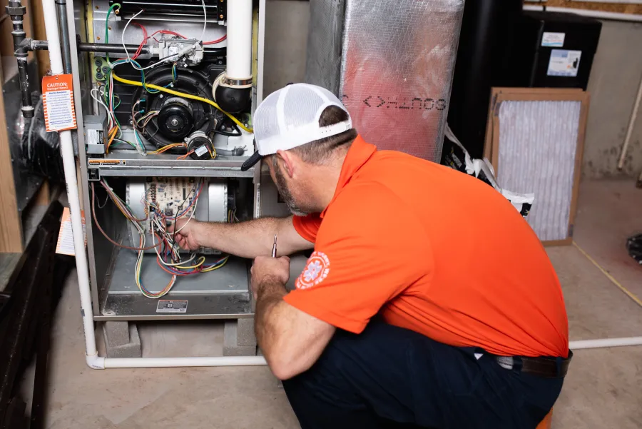 Technician in orange shirt inspecting and repairing wiring inside a residential furnace unit in basement