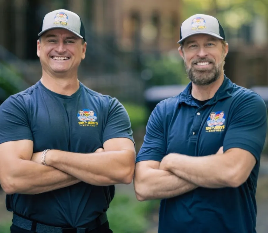 Two smiling men in matching blue shirts and hats with a roofing company logo stand outdoors with arms crossed.