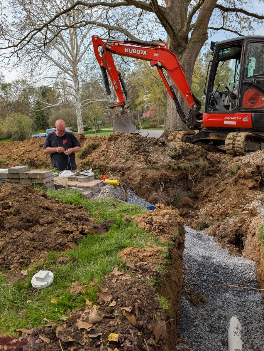 Man working near Kubota excavator digging trench for outdoor construction with gravel and pipes.