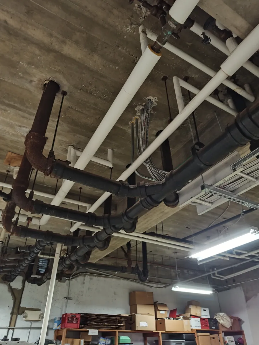 Ceiling view of industrial pipes and fluorescent lighting above storage shelves with boxes in a warehouse.
