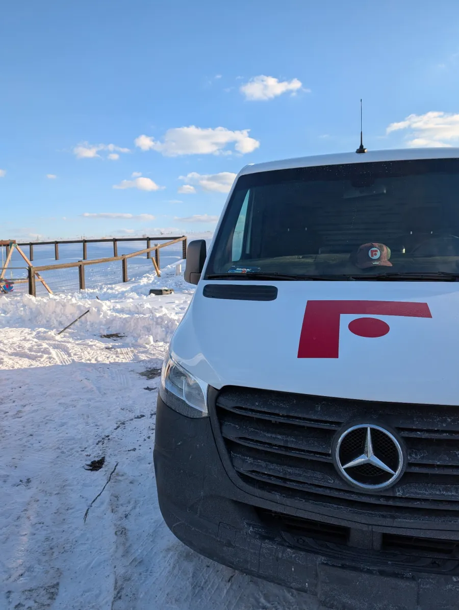 Front view of a white Mercedes van with a red logo parked on snowy ground under a blue sky with clouds.