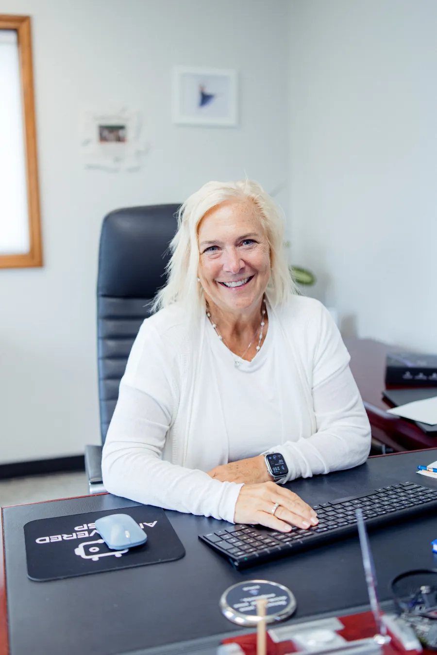 Smiling middle-aged woman with blonde hair sitting at office desk using keyboard with mouse on desk pad.