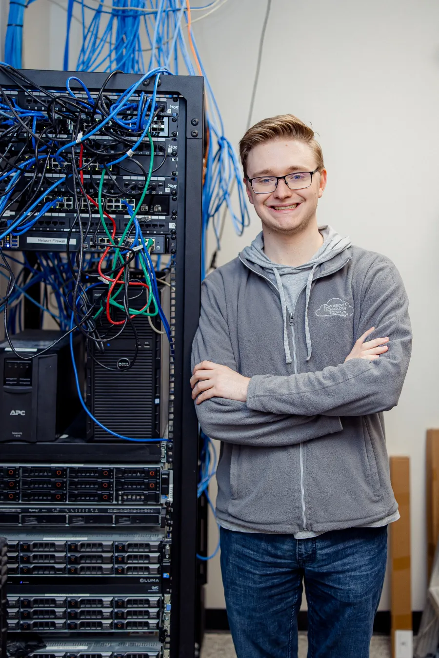 Young IT professional smiling with arms crossed next to server rack with cables and networking equipment.