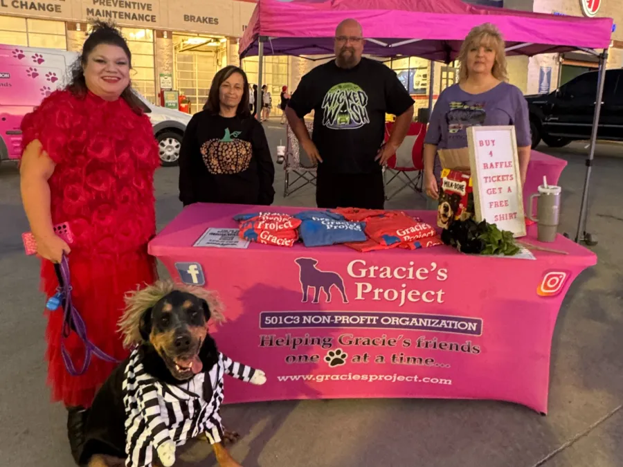 Volunteers at Gracie's Project booth with a dog dressed in a striped costume at an outdoor evening event.