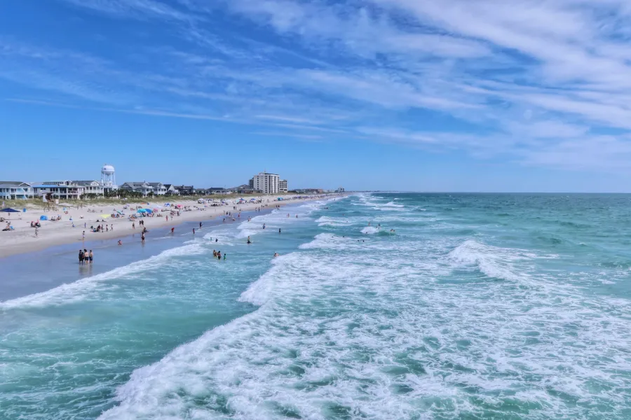 Wide sandy beach with waves, people swimming and relaxing under a blue sky with scattered clouds.