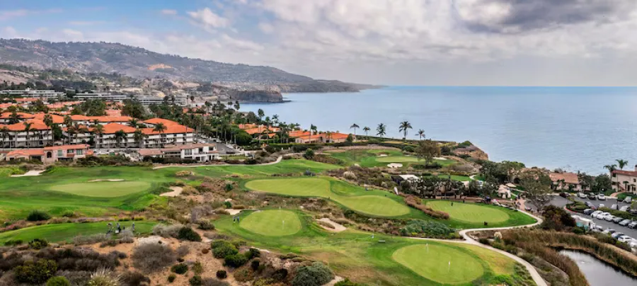 Rancho Palos Verdes Coastal golf course with green fairways, ocean view, and buildings with red-tiled roofs under cloudy sky.