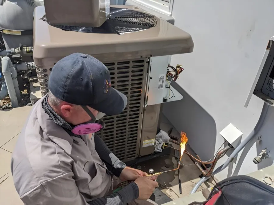 Technician wearing protective mask and cap soldering copper pipes near an HVAC unit outdoors on a sunny day