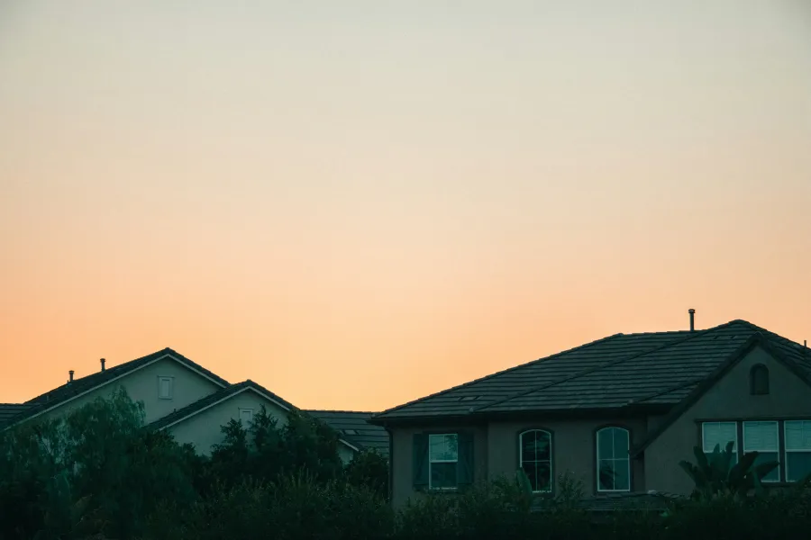Suburban houses with dark roofs against a clear orange sky at sunset, surrounded by greenery and trees.