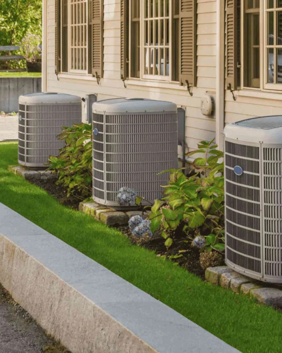 Three outdoor air conditioning units installed beside a house with green lawn and bushes.