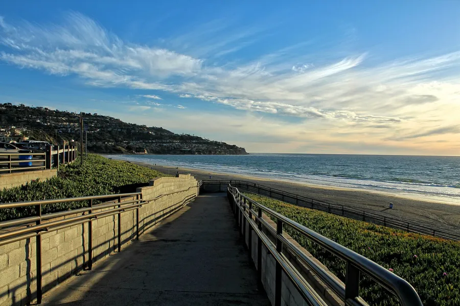 Pathway with railings leads to a sandy beach under a partly cloudy sky at sunset near a coastal hillside.