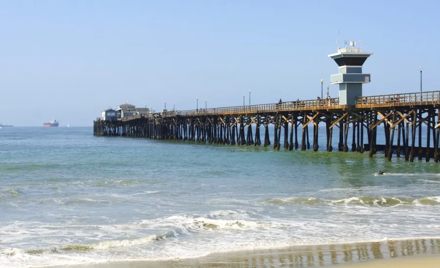 Wooden pier in Seal Beach, CA with a watchtower extending into calm ocean waters under clear blue sky at sandy beach