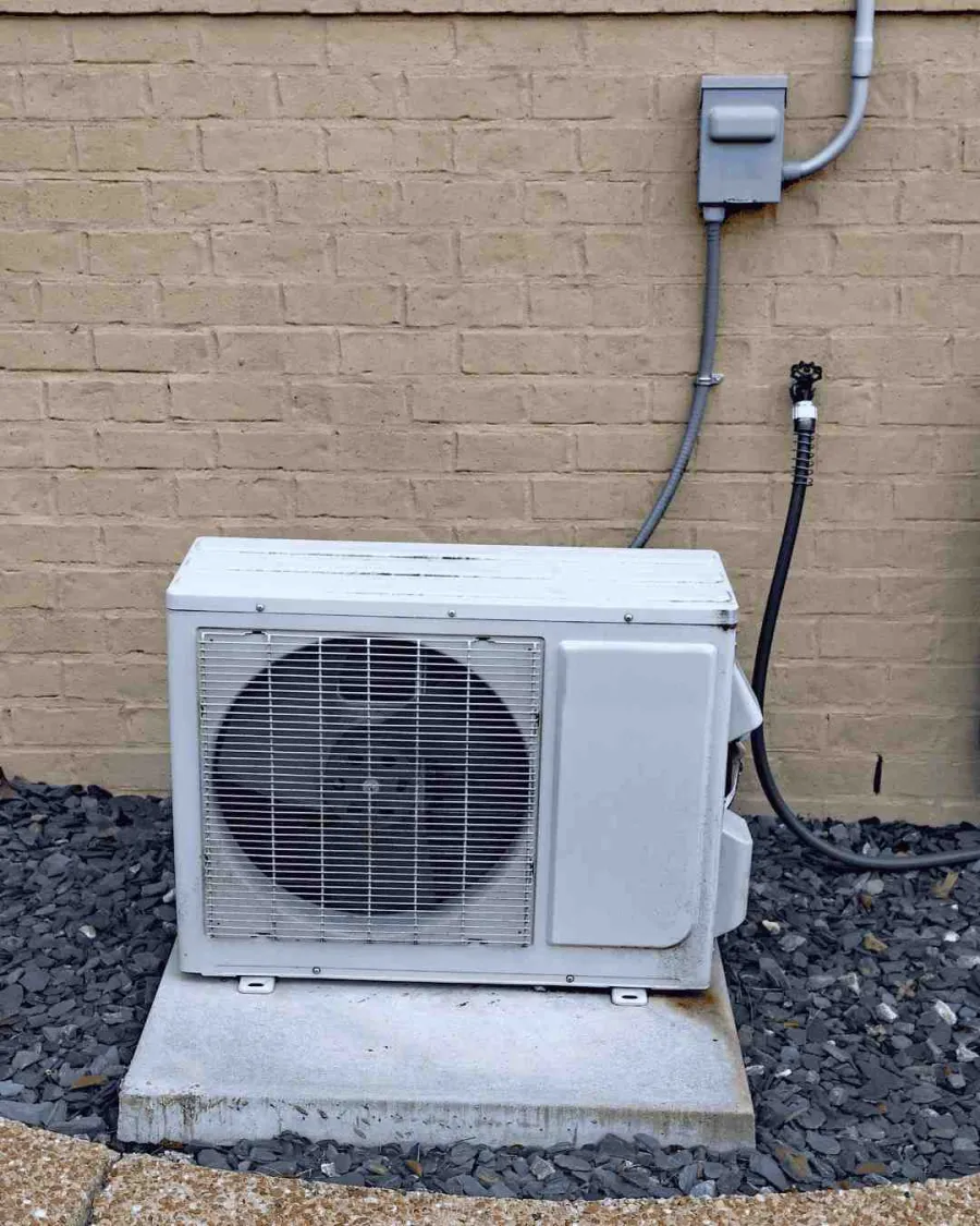 Outdoor air conditioning unit placed on concrete slab next to beige brick wall with electrical conduit.