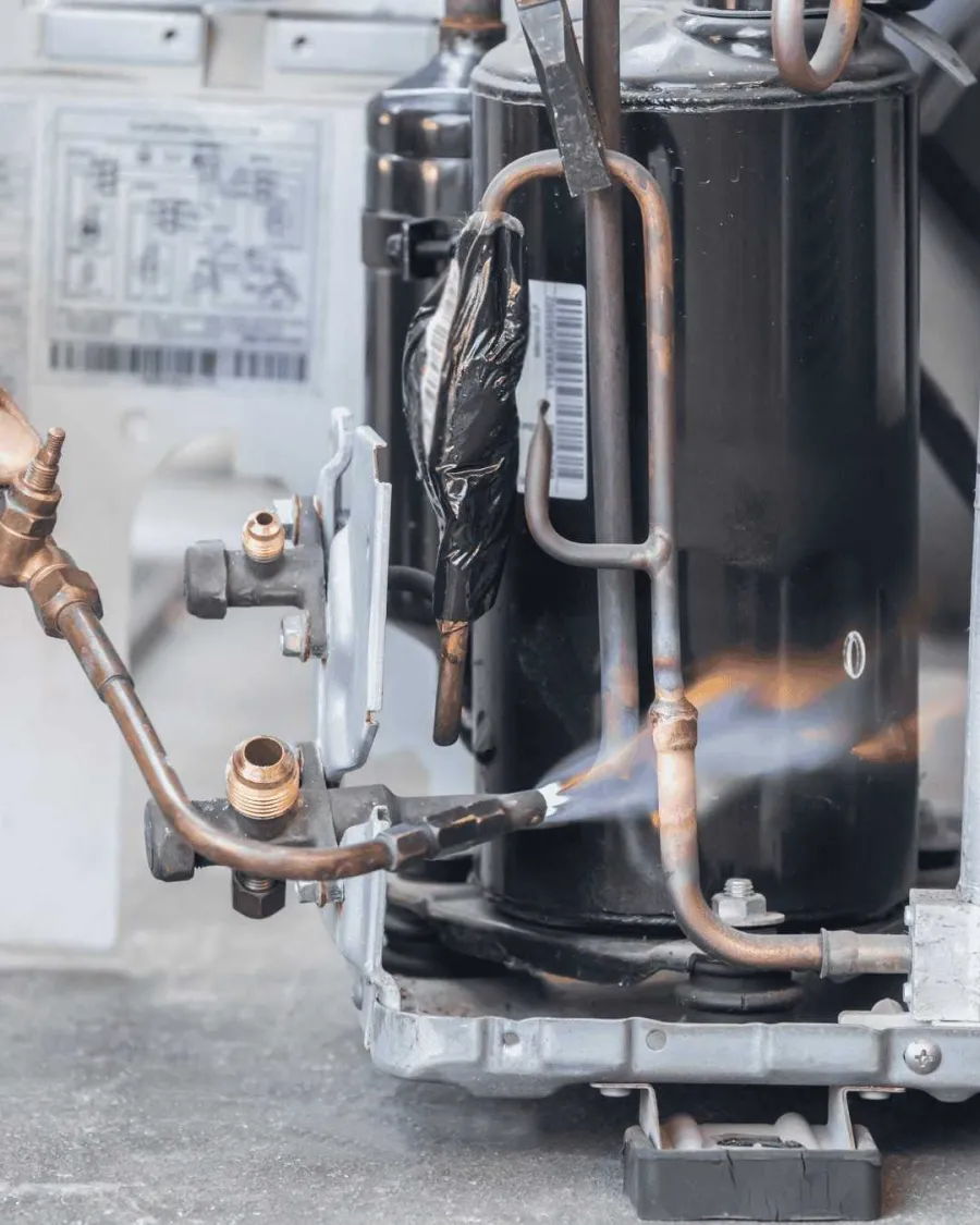 Close-up of technician using a flame torch to repair copper pipes on an HVAC compressor unit outdoors.