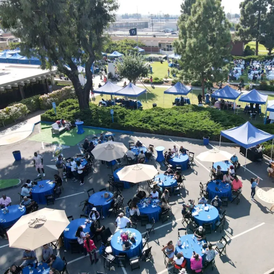 Outdoor community event with round blue tables, umbrellas, and people dining and socializing in a sunny park.