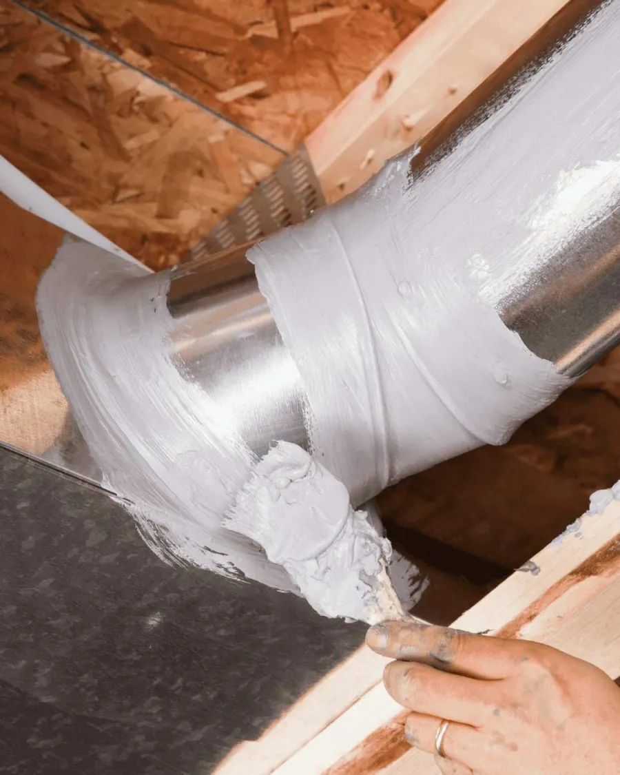 Person applying white sealant around metal duct pipe joint in wooden ceiling framework.