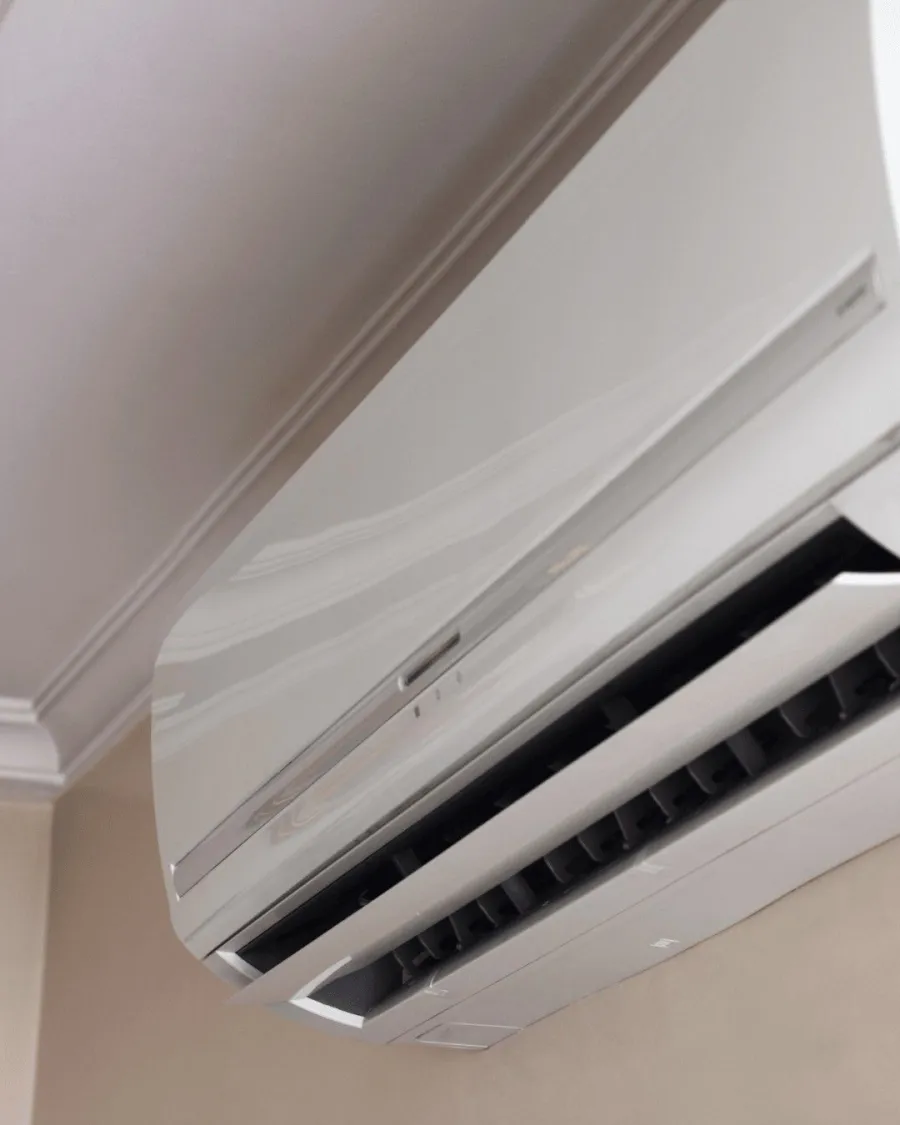 Modern white wall-mounted air conditioner unit installed near ceiling in a beige room corner.