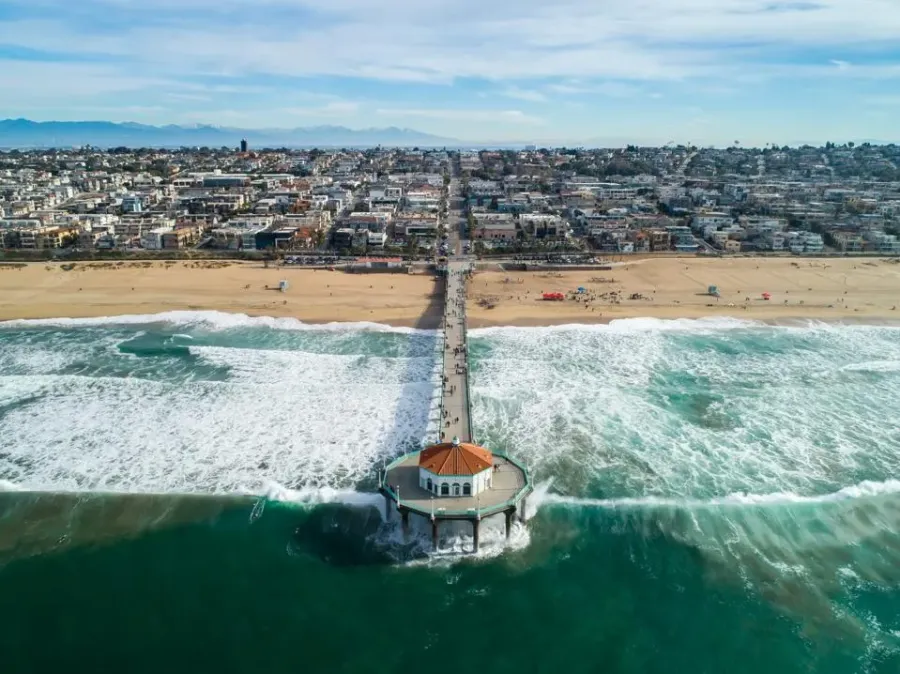 Aerial view of a coastal pier extending into the ocean with waves and a sandy beach in the background.