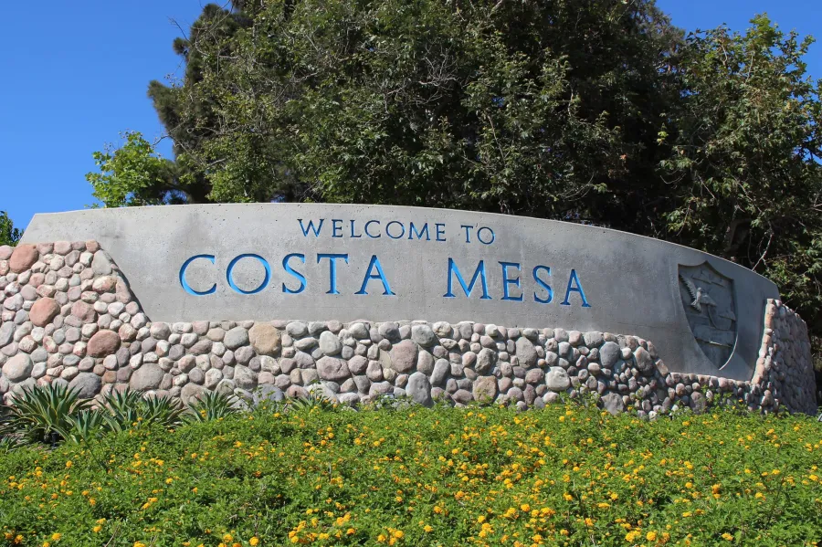 Welcome to Costa Mesa stone city sign with greenery and trees under clear blue sky.