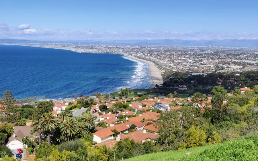 Palos Verdes Estates Coastal cityscape with red-roofed homes, palm trees, sandy beach, and blue ocean under a partly cloudy sky.
