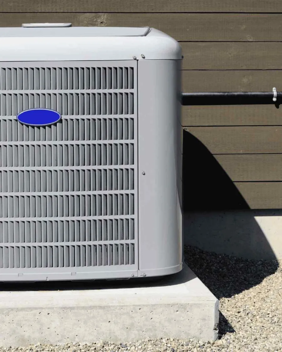 Gray outdoor air conditioning unit installed on concrete slab next to a wood paneled house wall.