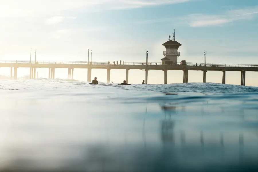 Ocean view with surfers near a pier and lifeguard tower under a clear blue sky at sunrise.