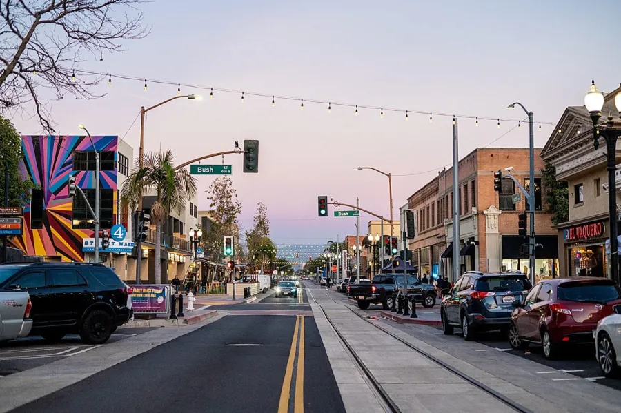 Downtown street in Santa Ana, CA scene at dusk with cars, colorful mural, streetlights, and shops along both sides of the road.