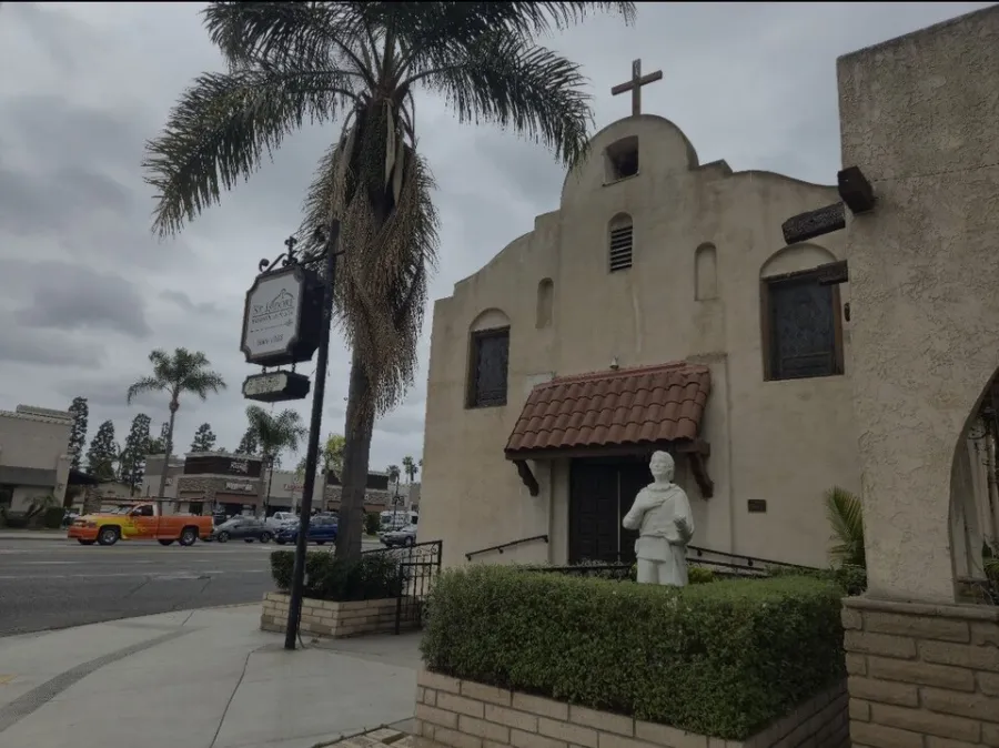Historic beige church with a cross on top, statue in front, and palm trees on a cloudy day in urban area.
