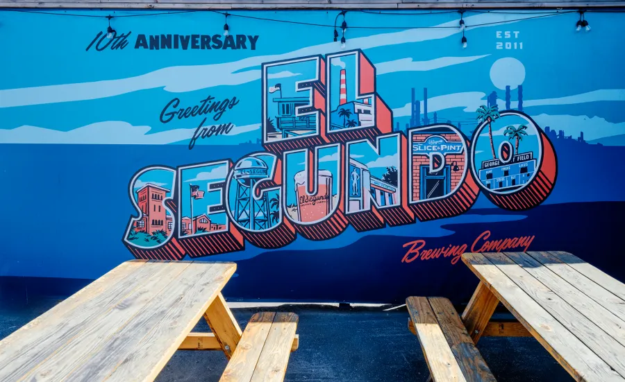 Colorful mural celebrating El Segundo Brewing Company's 10th anniversary on blue wall behind wooden picnic tables.