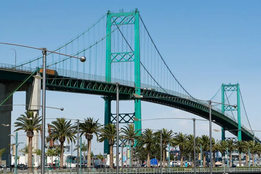 Green suspension bridge in San Pedro, CA over palm trees and urban street under clear blue sky.
