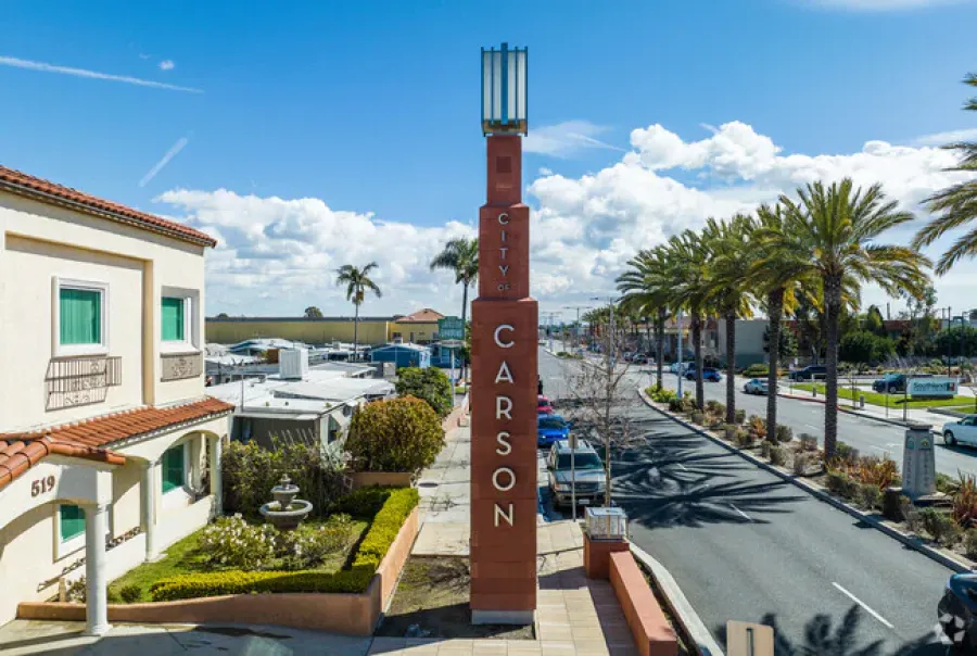 City of Carson pillar sign beside street lined with palm trees and buildings under a blue sky