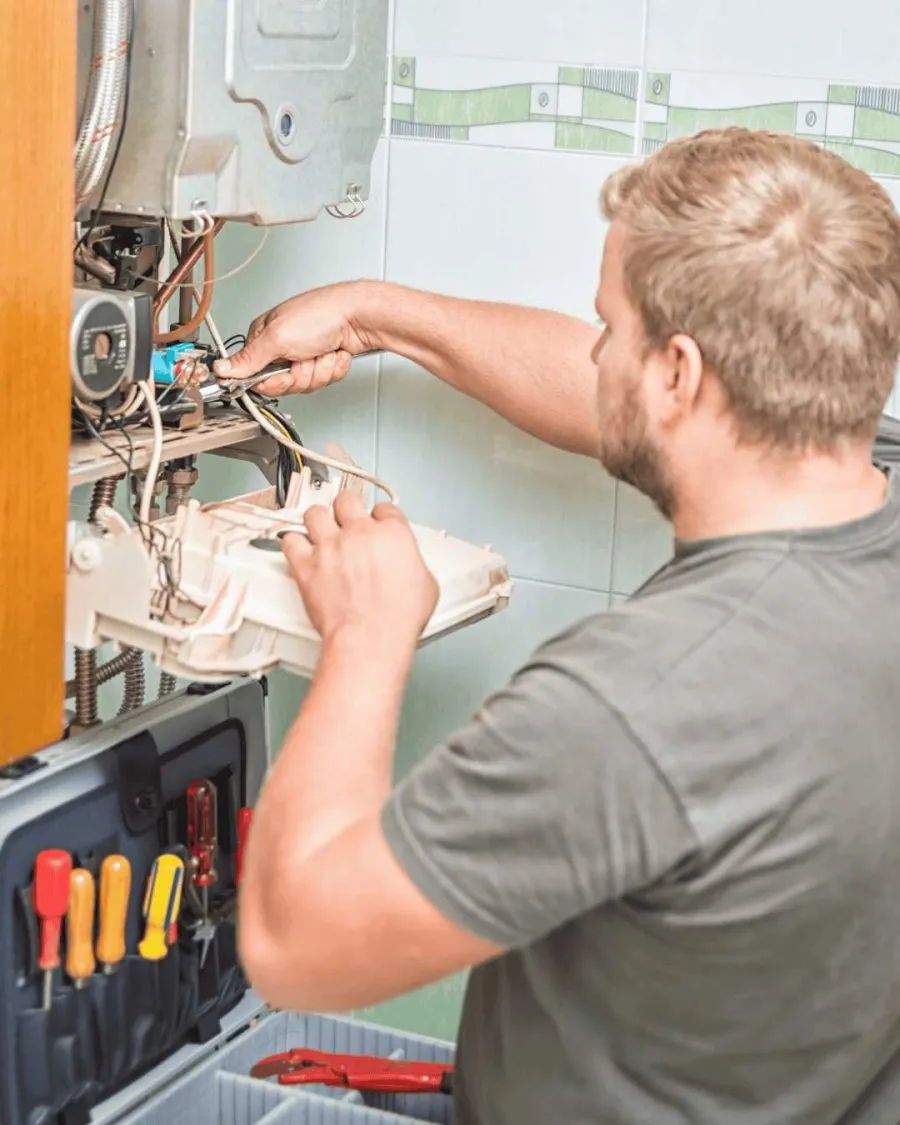 Technician repairing a water heater using tools from an open toolbox in a tiled indoor space.