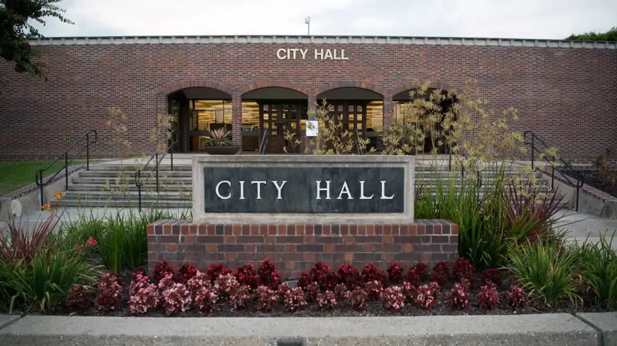 Brick city hall building with steps, flower beds, and a large City Hall sign in front on a cloudy day