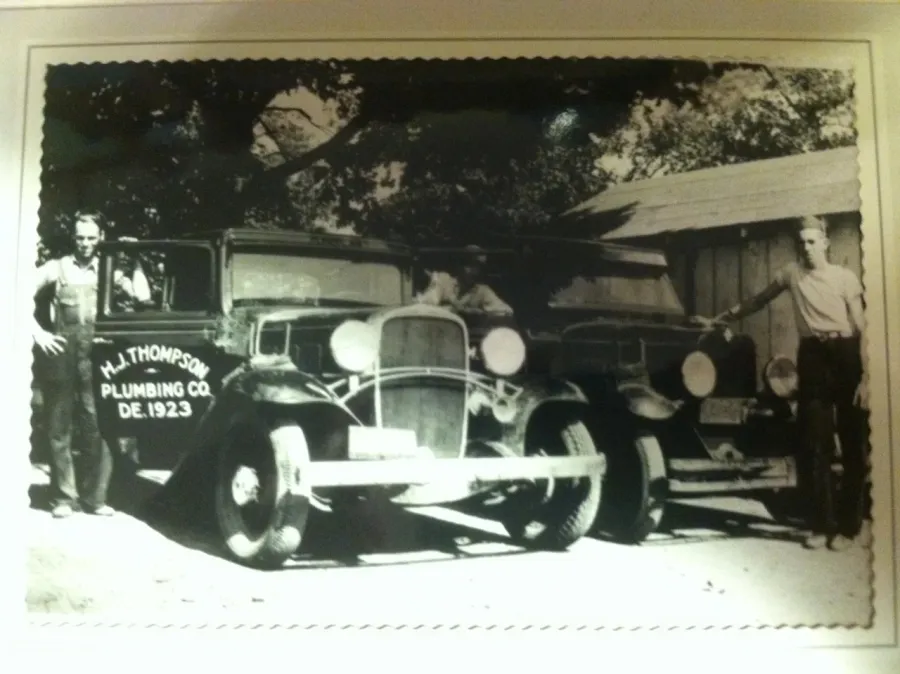 Black and white photo of two men standing beside vintage cars, one marked H.J. Thompson Plumbing Co. 1923.